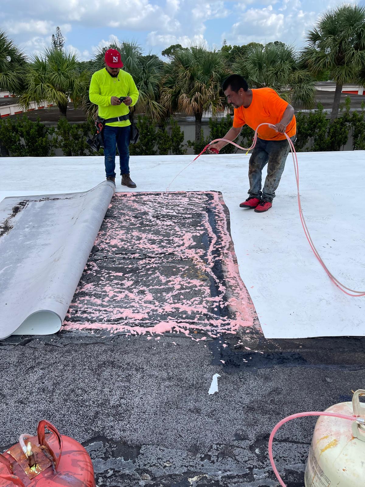 Two workers repair a flat roof, peeling back material and using equipment with pink adhesive foam.