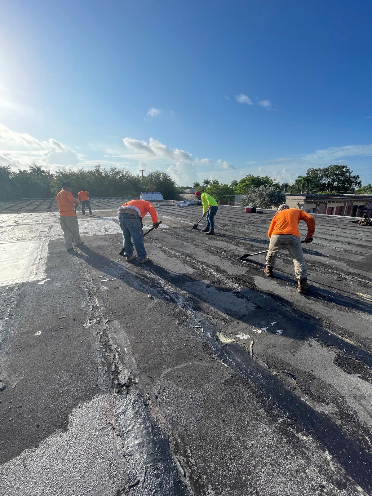Four workers in bright shirts repair a flat rooftop under a clear blue sky with scattered clouds.