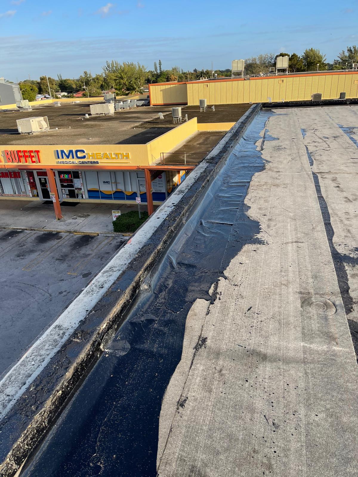View of a flat rooftop with damaged roofing material above a strip mall with stores and trees in the background.