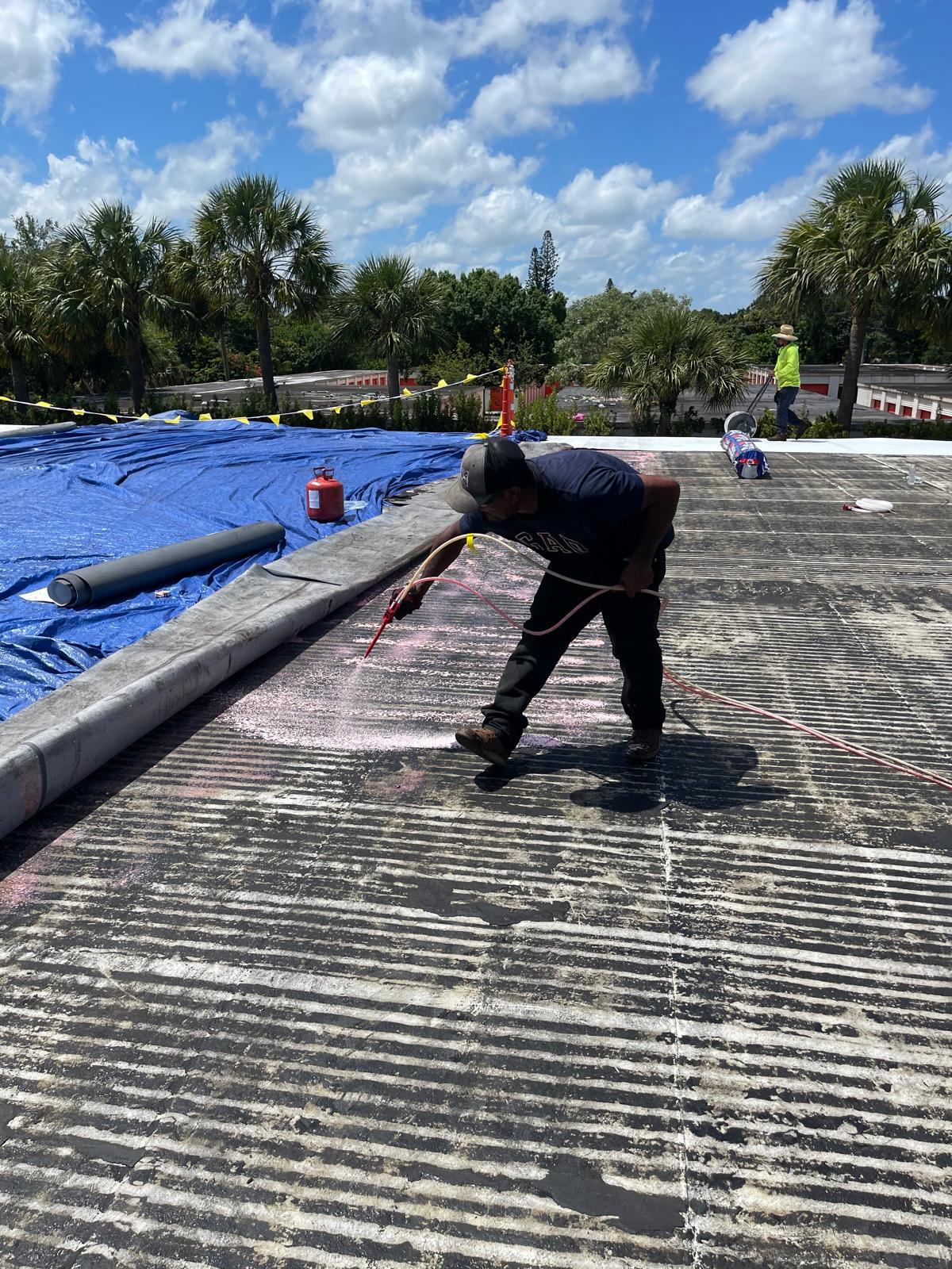 A worker uses a tool on a rooftop near a blue tarp, with palm trees and a cloudy sky in the background.