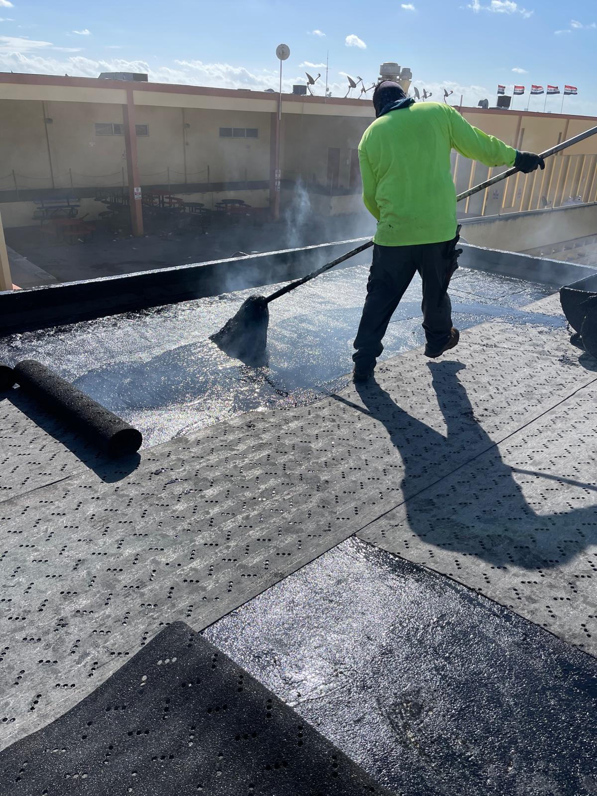 Worker in a neon shirt applies roofing material with a roller on a flat roof under bright sunlight.