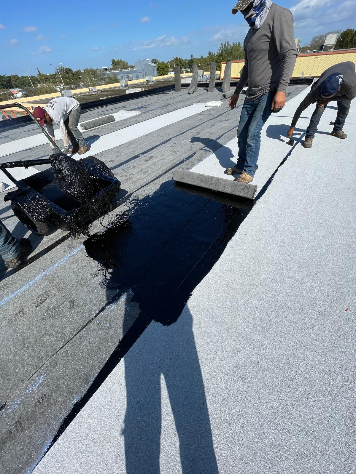 Workers installing roofing materials and spreading black tar on a flat rooftop under a sunny sky.