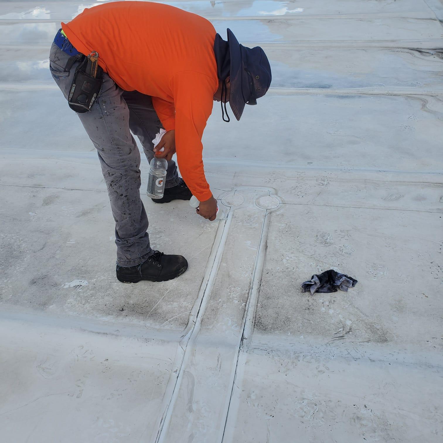 Worker in orange shirt sealing seams on a white roof with a tool and bottle, rag nearby.