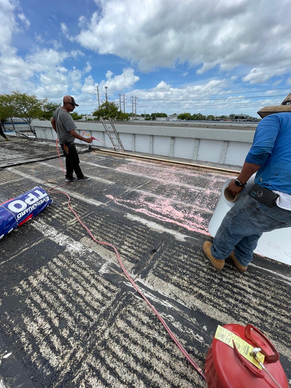 Two workers on a rooftop apply materials with tools under a partly cloudy sky, preparing the roof surface.