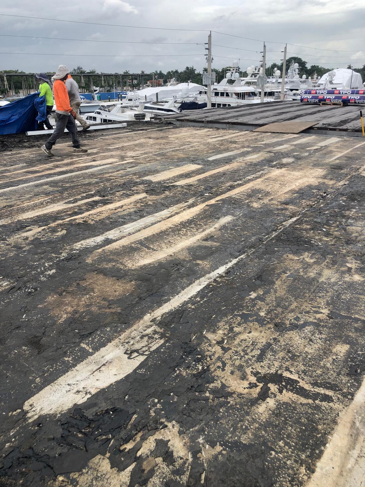Two construction workers walk on a weathered rooftop near a marina with several docked boats in the background.