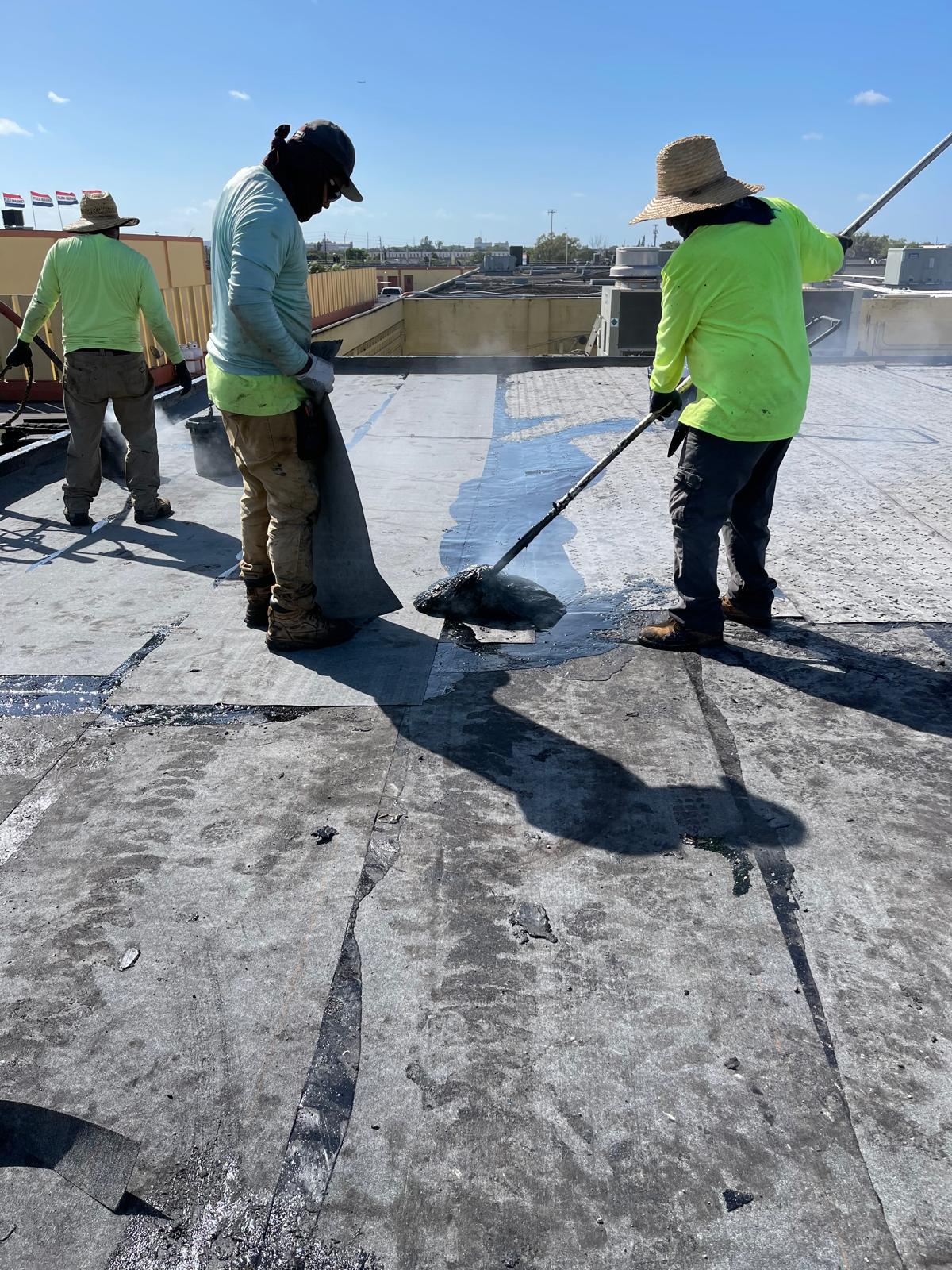 Two workers in hats and neon shirts install roofing material under bright sunlight on a flat rooftop.