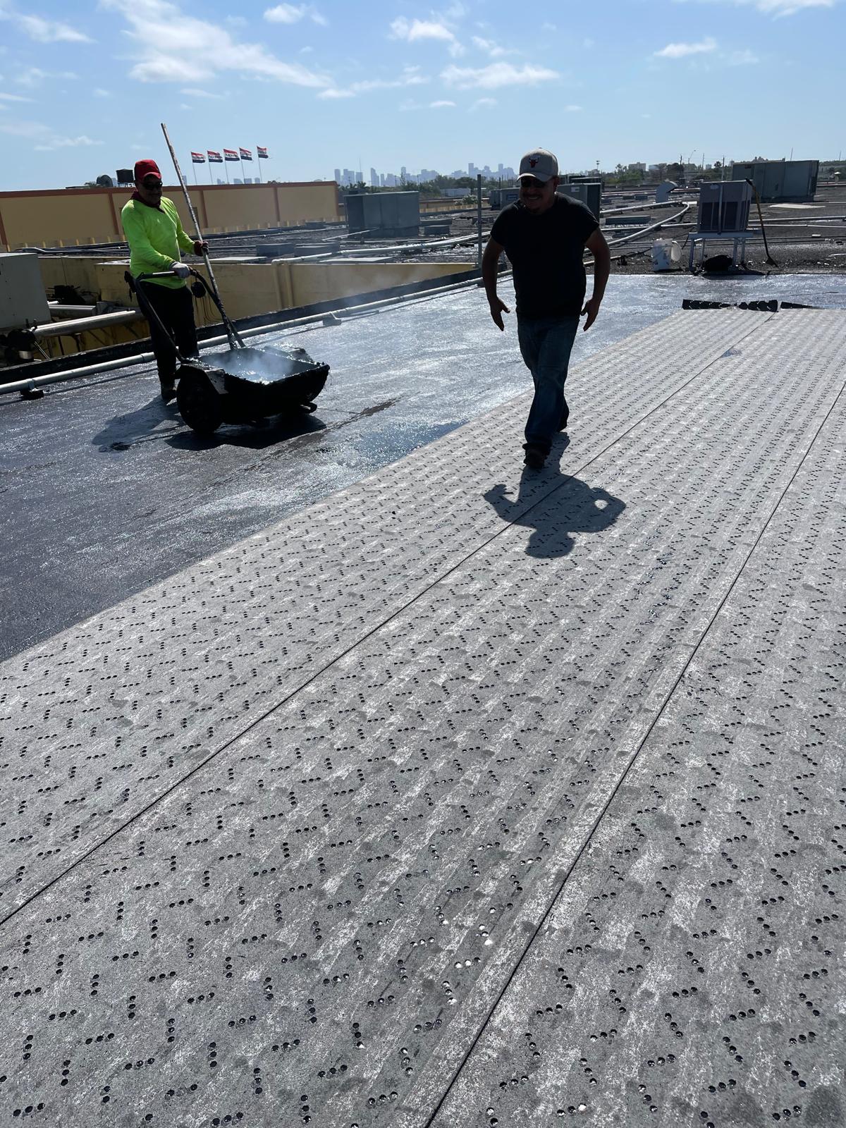 Two workers apply roofing material and hot tar on a flat rooftop under a sunny sky.