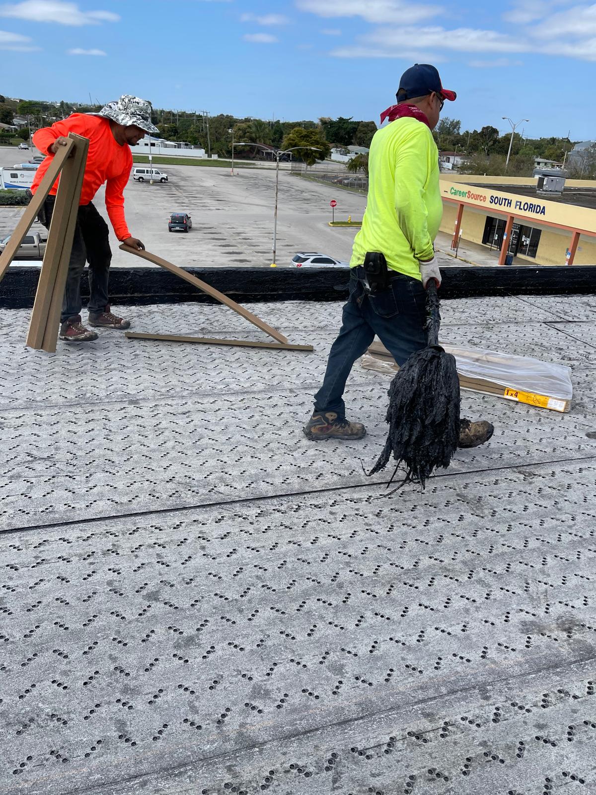 Two workers on a rooftop carry tools and materials, with a shopping plaza visible in the background.