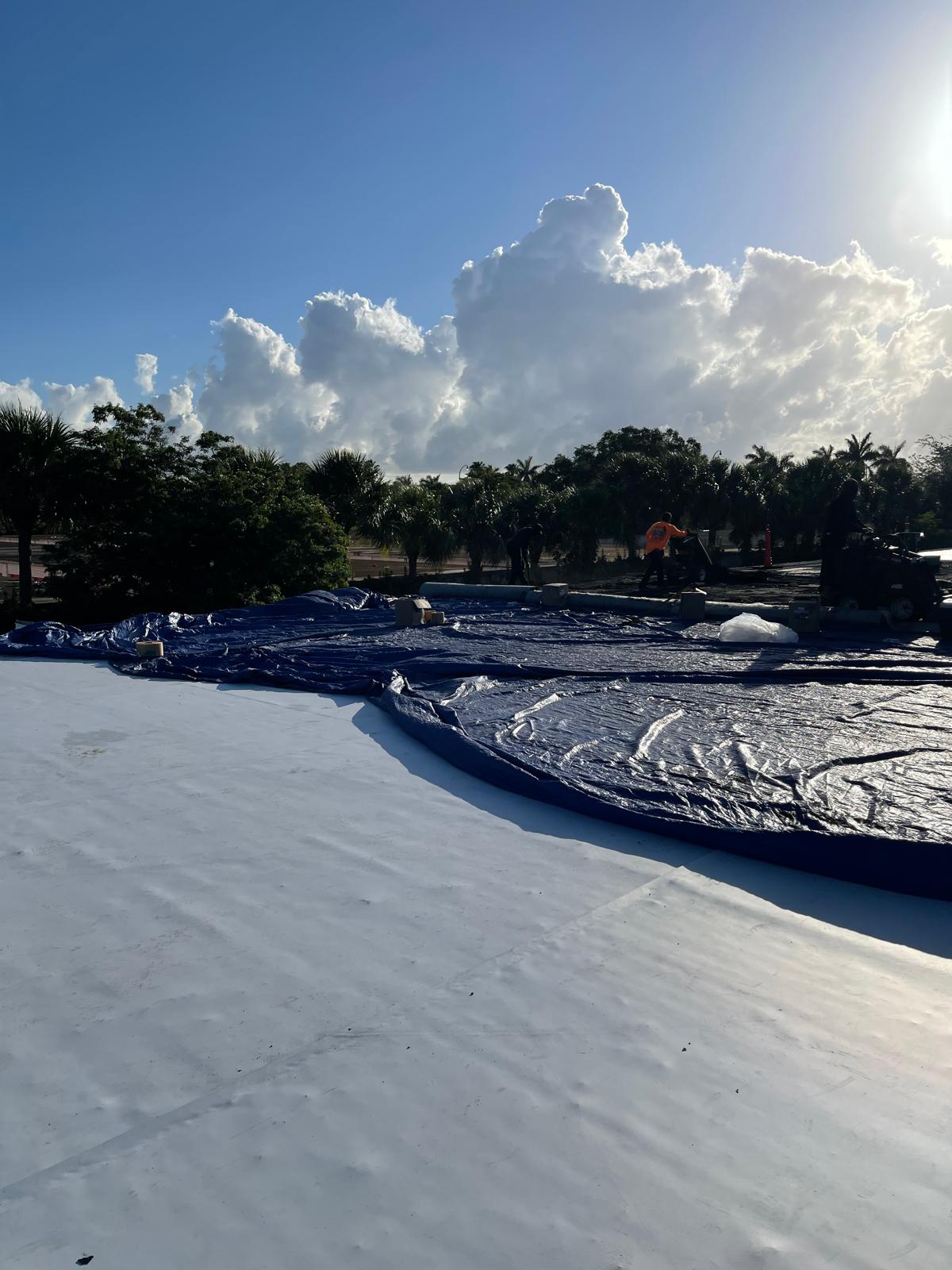 Workers with tarps on a rooftop under a cloudy sky; trees and bright sunlight in the background.
