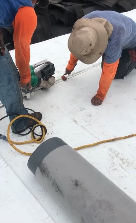 Two workers in hats working on a white roof with tools and a yellow extension cord.