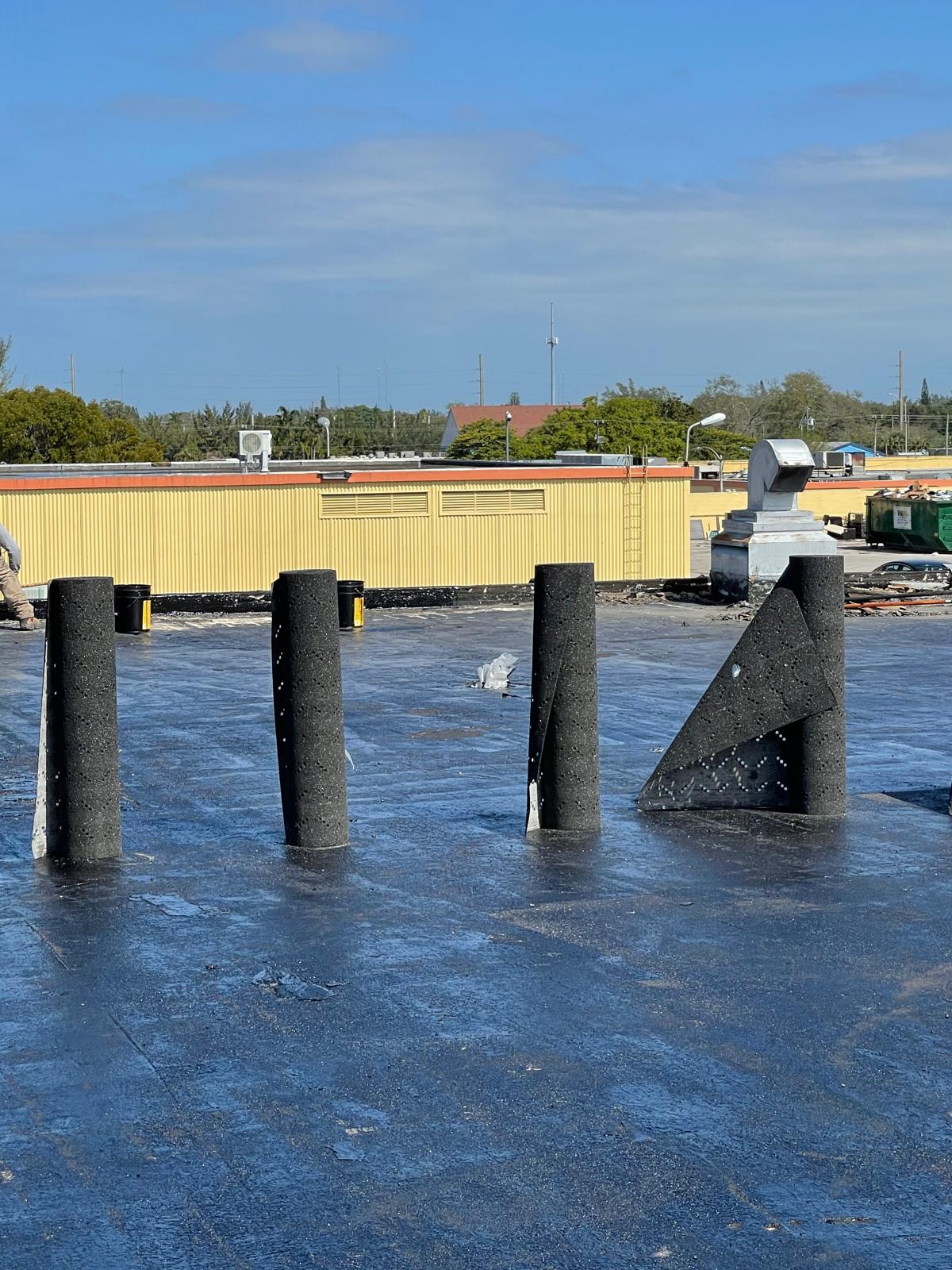 Four rolls of roofing material stand upright on a flat, black roof under a clear blue sky.