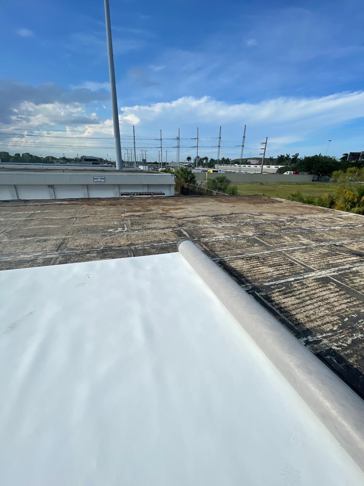 A rooftop with a section of new white material being installed, overlooking a field and power lines.