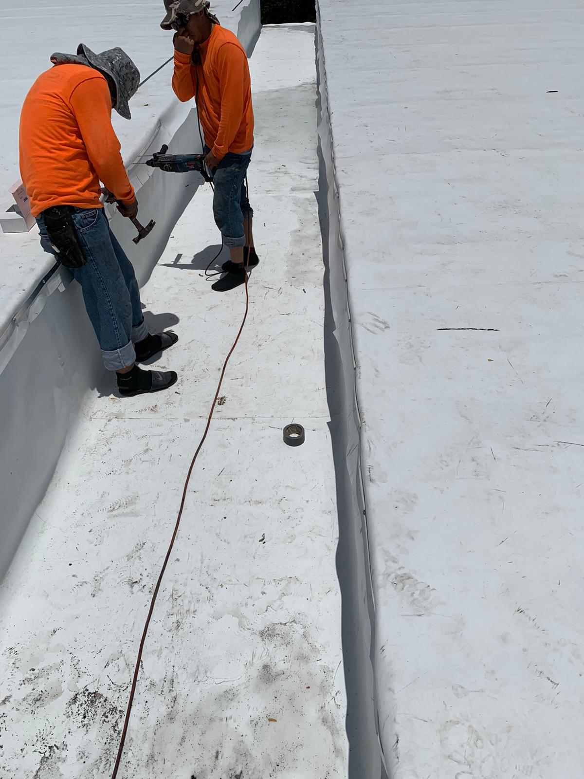 Two workers in orange shirts working on a white rooftop with tools and a long cable between them.