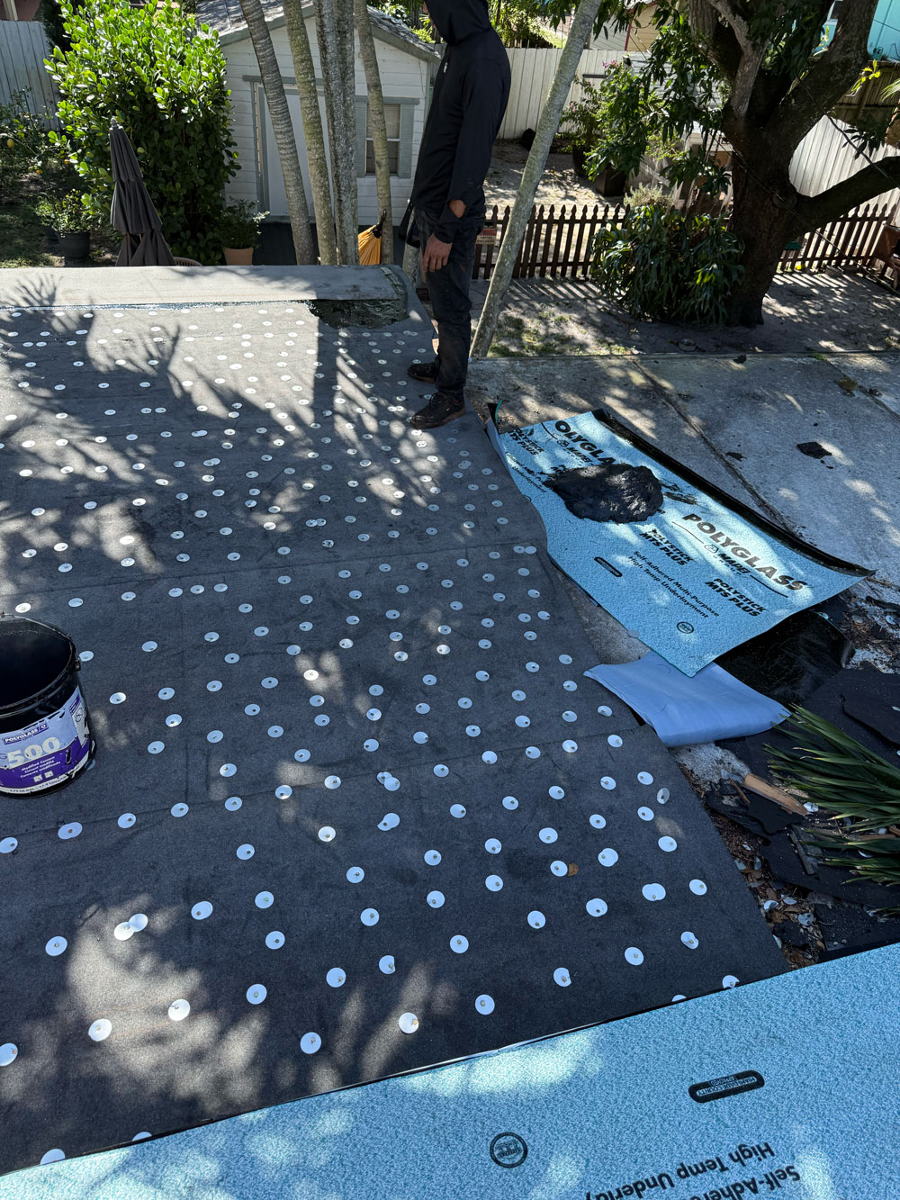 A person stands on a roof under repair, with roofing materials and tools scattered around.