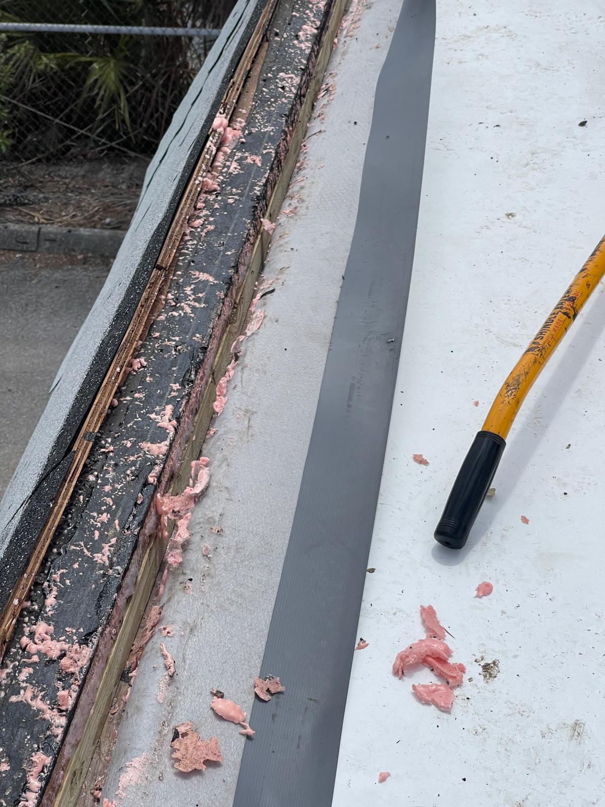 Close-up of a roof edge with pink insulation, a metal strip, and a yellow-handled tool lying on the surface.