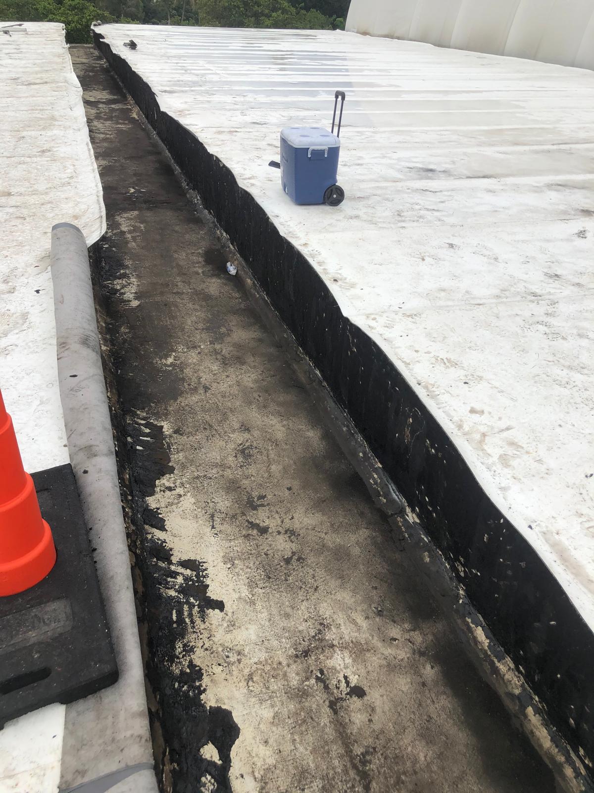 A blue cooler on wheels sits on a white, flat rooftop near a blackened drainage channel and orange safety cone.