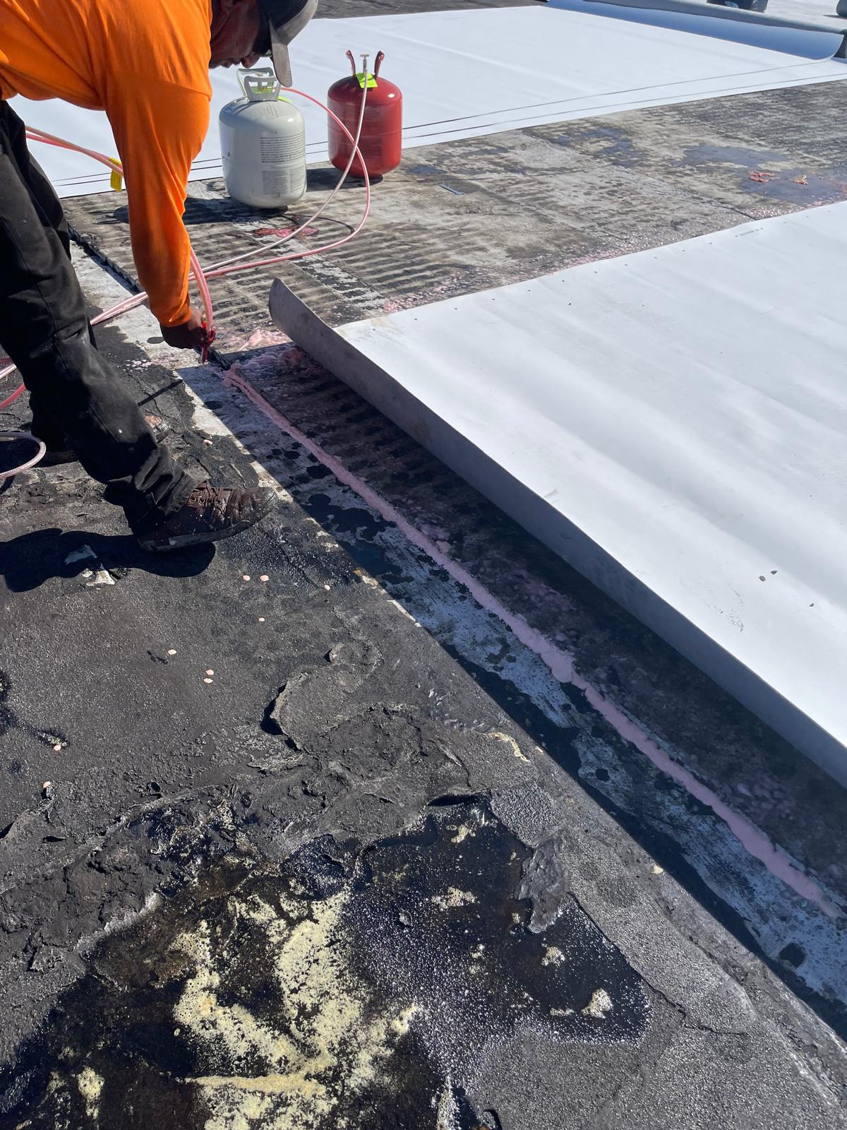 A worker installs white roofing insulation panels on a weathered flat roof, with gas tanks nearby.