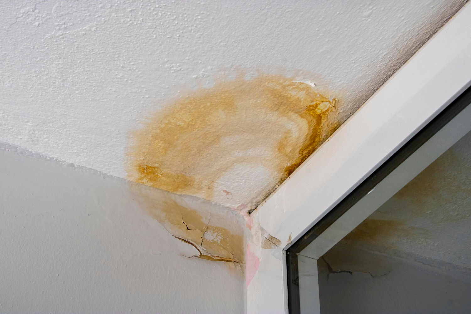 352 Brown water stain and cracks on a white ceiling near a corner by a window, indicating possible water damage.