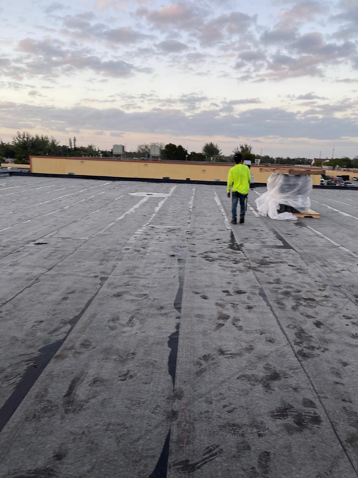 Person in a neon shirt stands on a flat rooftop covered in footprints, near wrapped materials, at sunset.