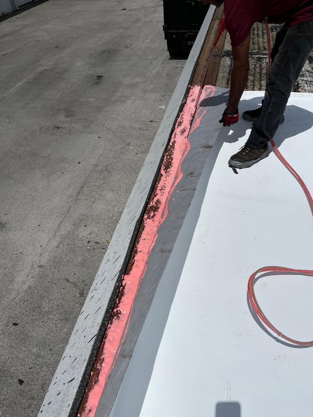 Person applying pink sealant along the edge of a roof with a hose on a sunny day.