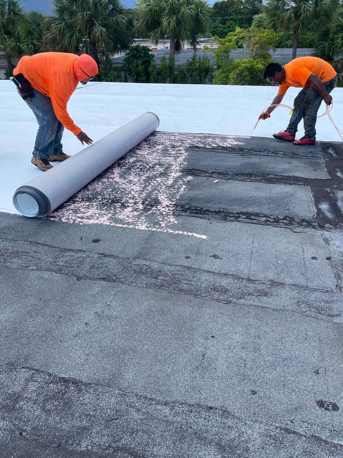 Two workers in orange shirts repair a flat roof, rolling out new material over an old surface.