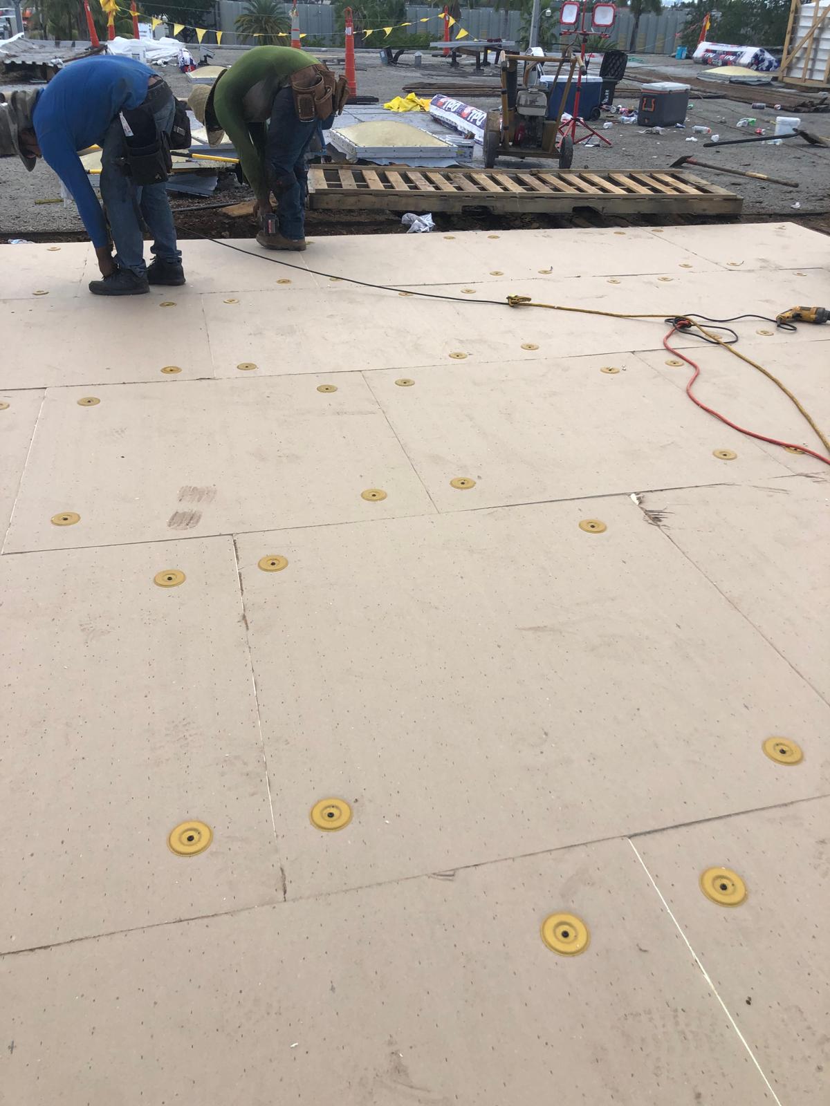 Two workers install insulation boards on a flat roof, with tools and materials scattered around.