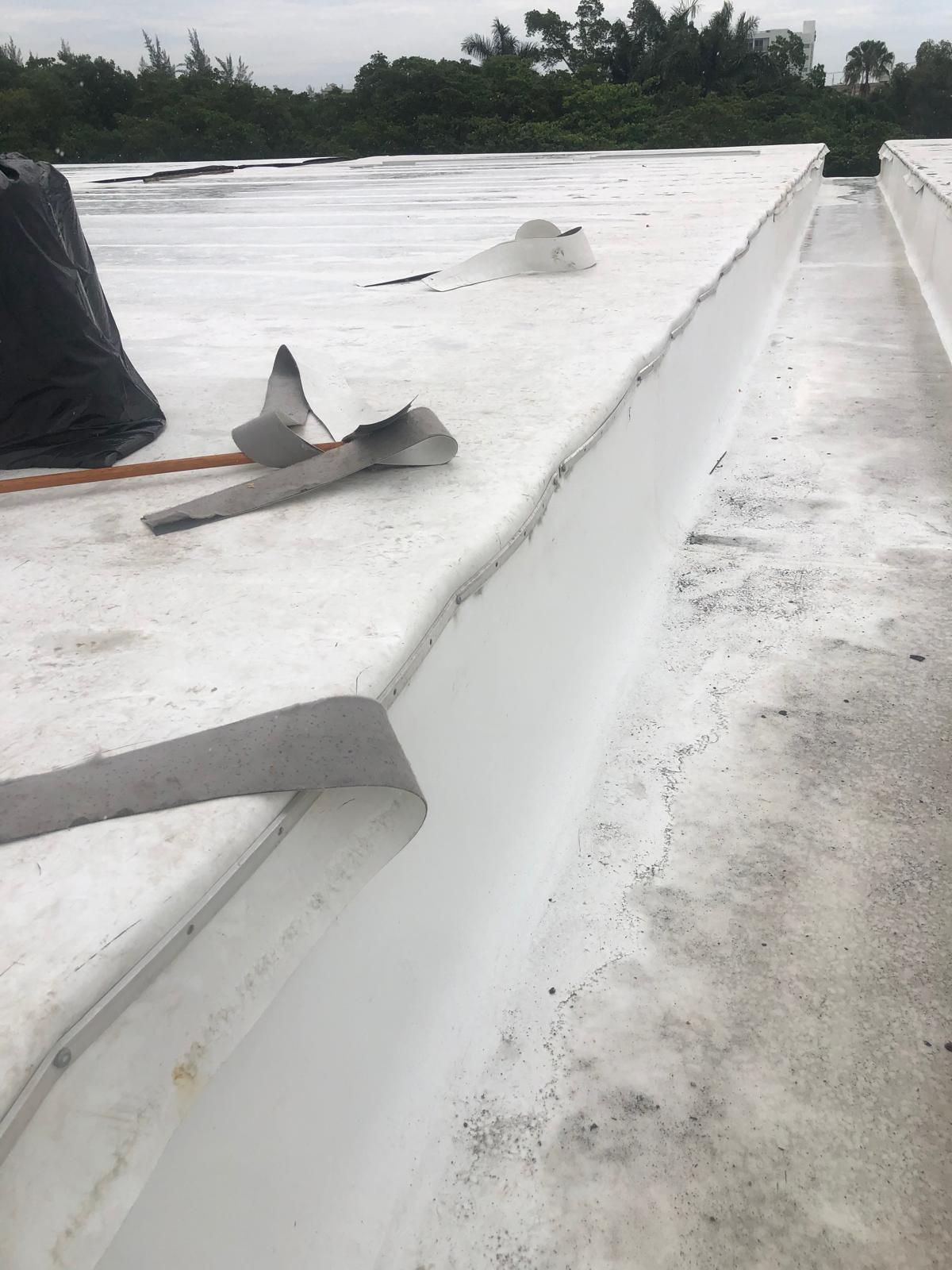 White flat roof with loose strips of roofing material and a black bag on a cloudy day, surrounded by greenery.