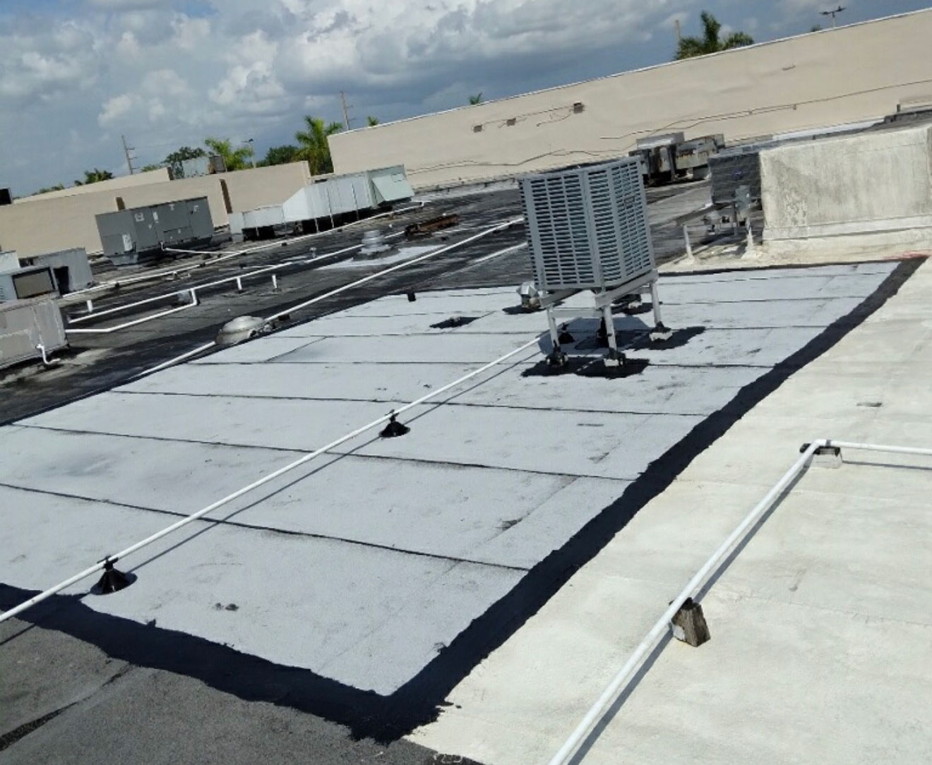 Rooftop with HVAC units and pipes, freshly coated section visible under a partly cloudy sky.