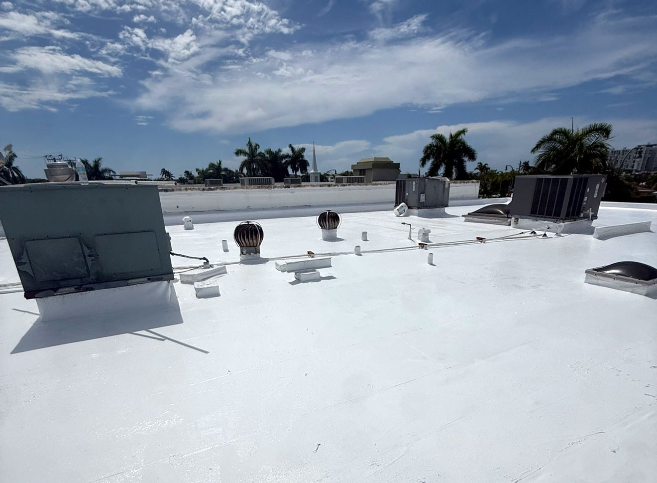 White rooftop with various HVAC units and vents, palm trees, and a partly cloudy blue sky in the background.