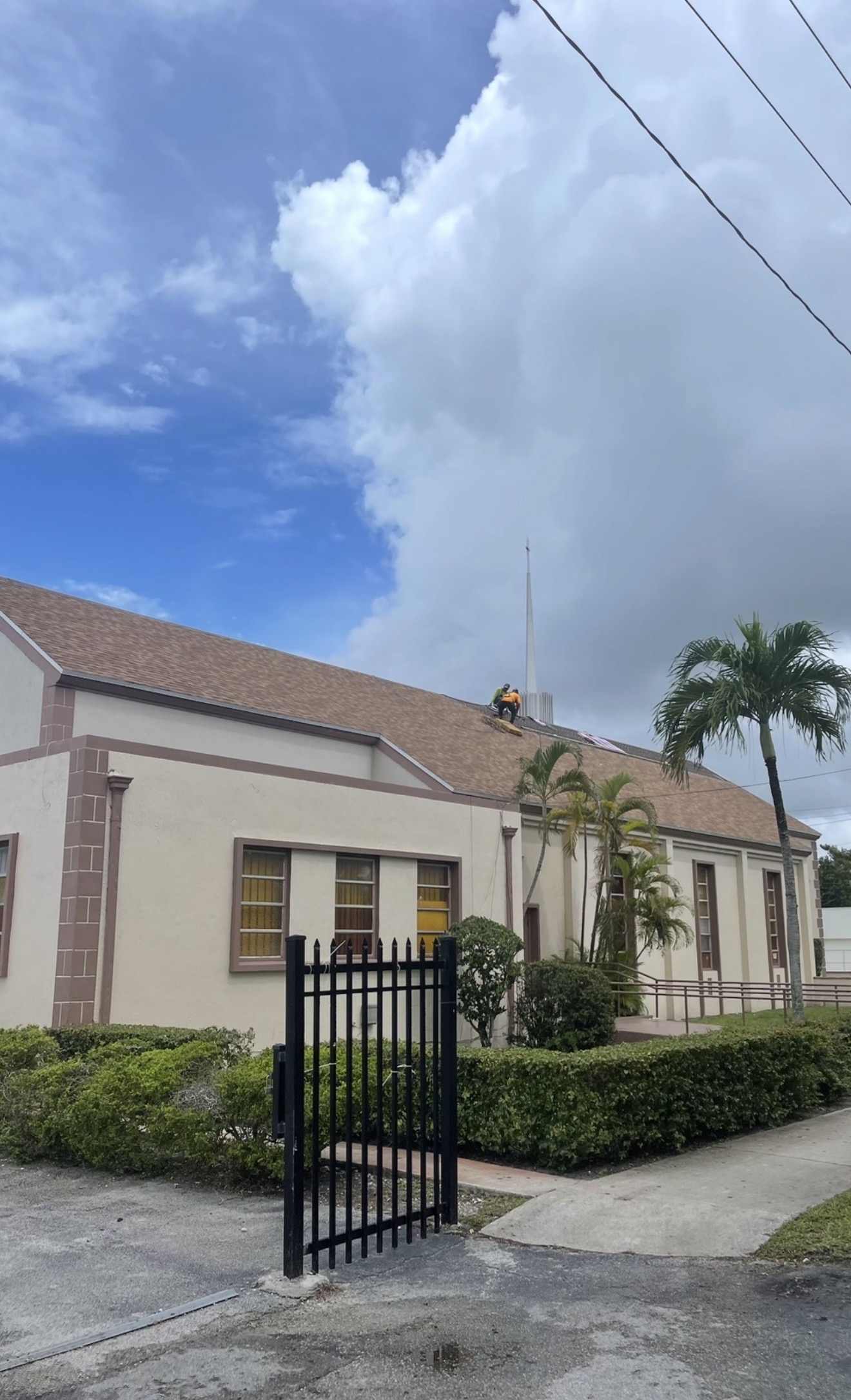 A person in a safety vest works on the roof of a beige church with palm trees and a black gate nearby.