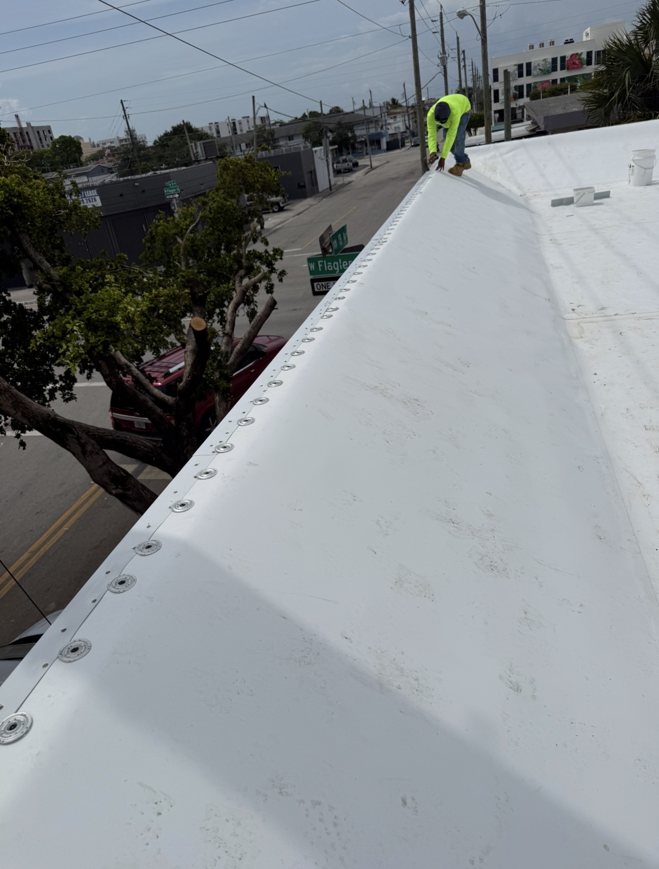 Worker in neon shirt on a white rooftop near bolts, trees, and street signs along a city road.