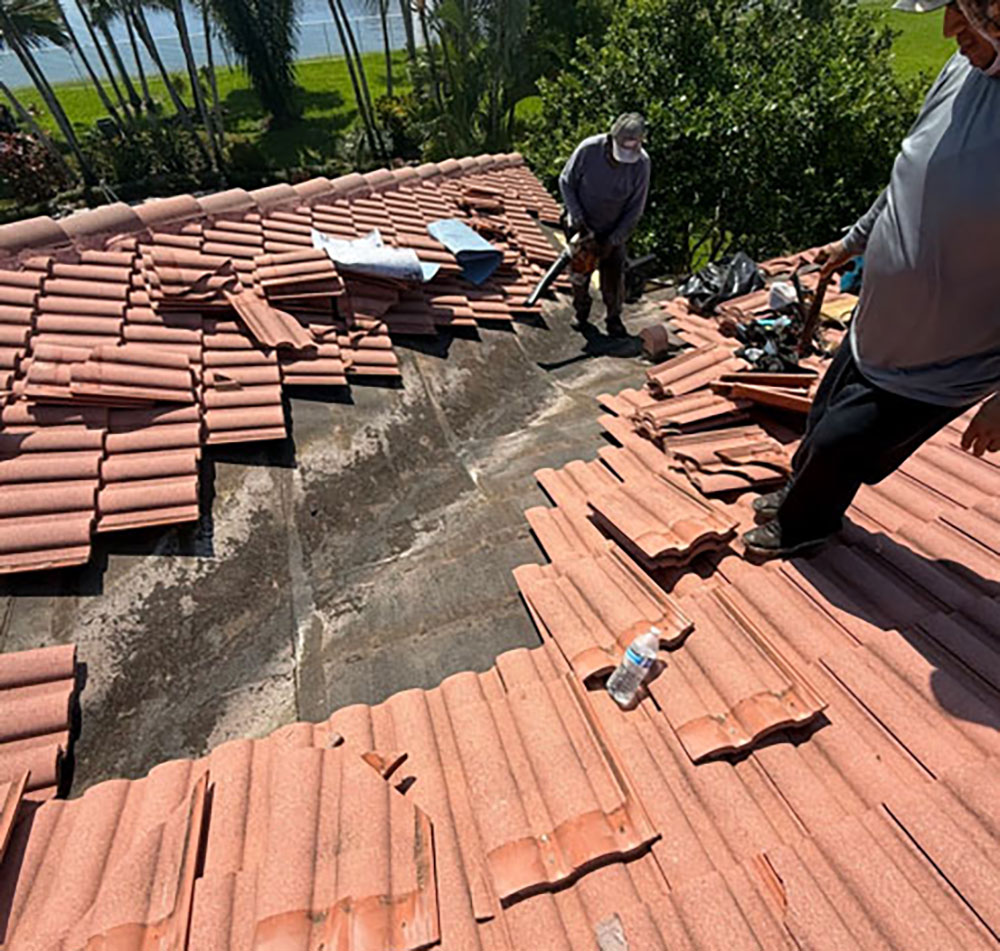 Two workers repair a roof with red tiles, some tiles removed, tools and a water bottle are visible.