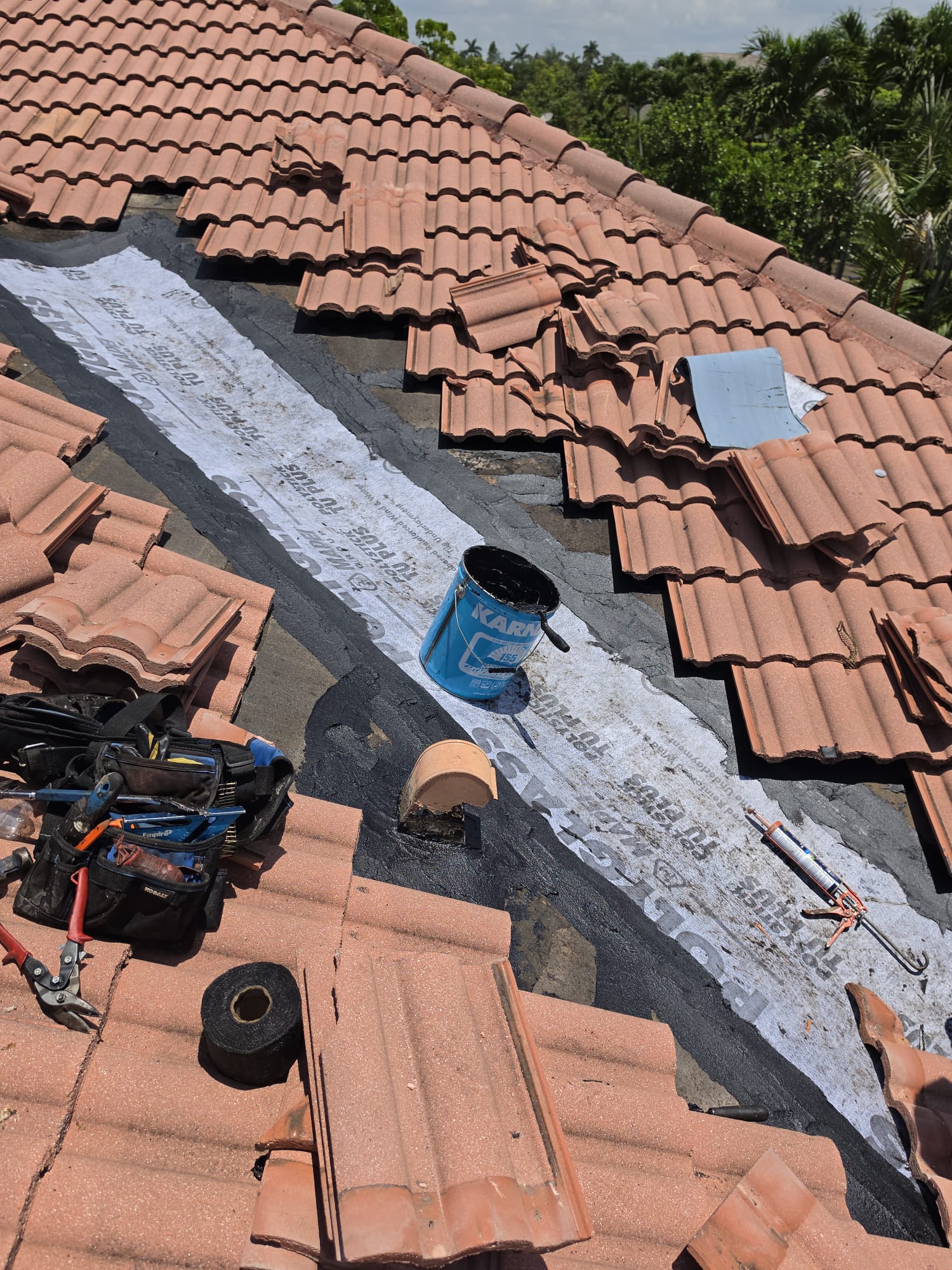 Roof repair in progress with tools, tiles, sealant, and a bucket placed on exposed underlayment.