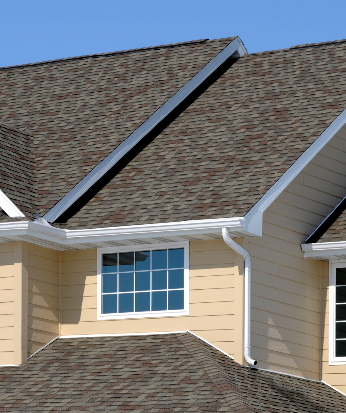 A beige house with a window, white trim, and brown shingle roof against a clear blue sky.