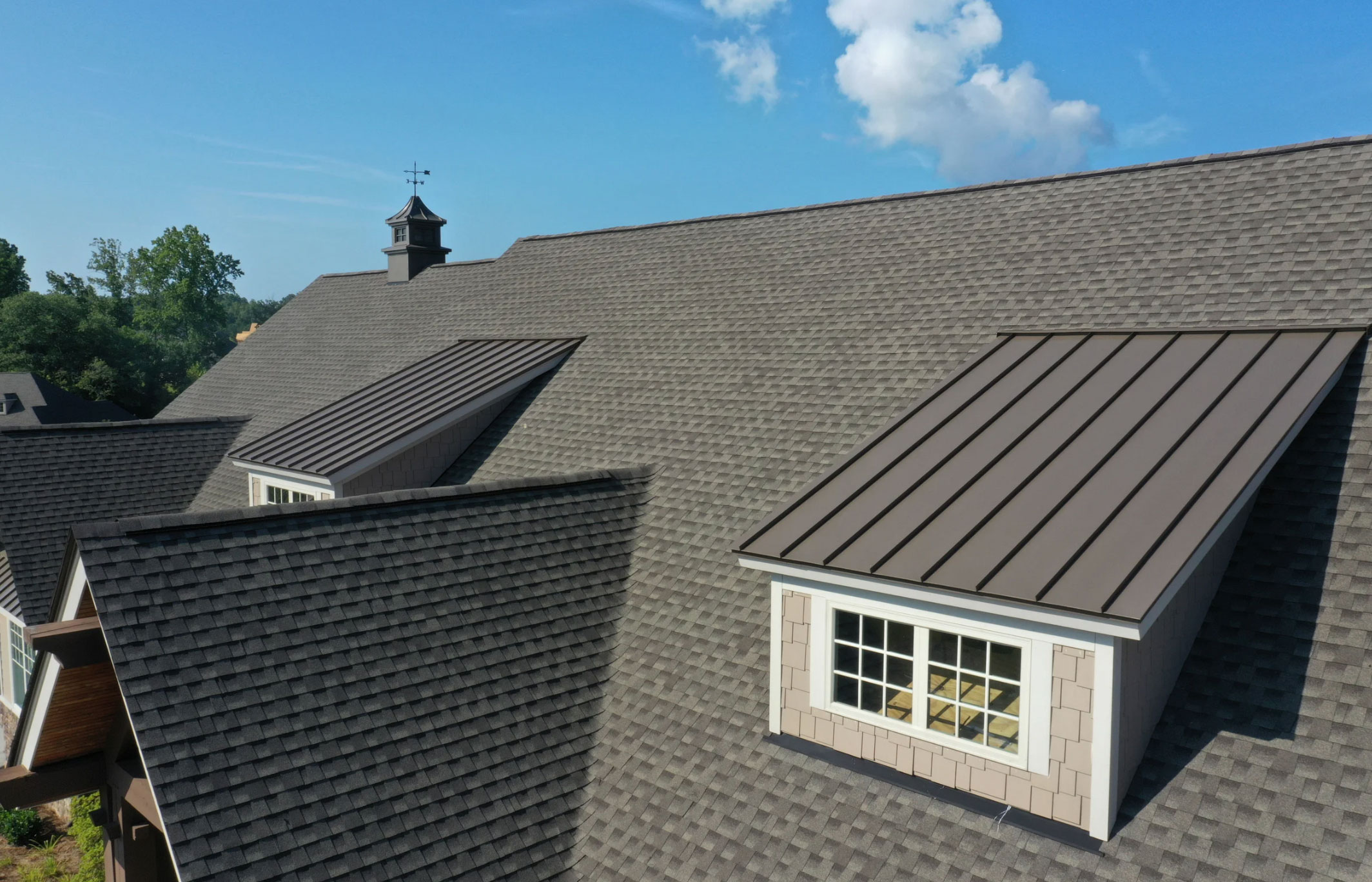 Close-up of a house roof with dormer windows and brown shingles under a blue sky with some clouds.