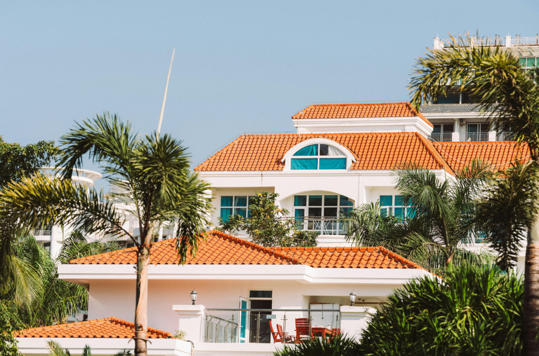 A white house with orange tile roof and blue windows, surrounded by palm trees under a clear sky.